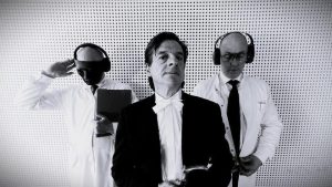 Die Privatiers Bandfoto. Im Vordergrund der Sänger im Frack, dahinter Gitarrist und Schlagzeuger im Arztkittel mit Kopfhörer und Maske. Das Trio steht vor einer weißen Lochrasterwand.